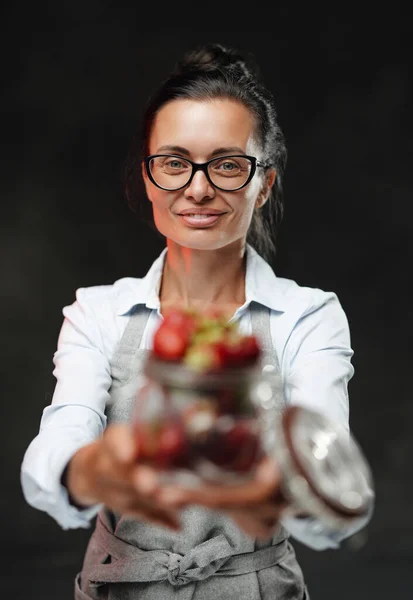 Bella donna di mezza età tiene un bicchiere con bacche succose rosse fresche. Studio foto su sfondo scuro — Foto Stock