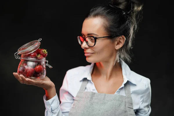 Bella donna di mezza età tiene un bicchiere con bacche succose rosse fresche. Studio foto su sfondo scuro — Foto Stock