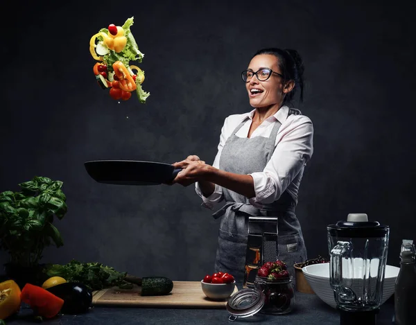 美しい成熟した女性キッチンで野菜サラダを調理し、パンから野菜をトス — ストック写真