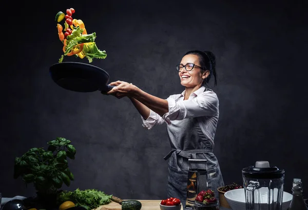 Chef feminino de meia-idade feliz jogando legumes cortados no ar de uma frigideira — Fotografia de Stock