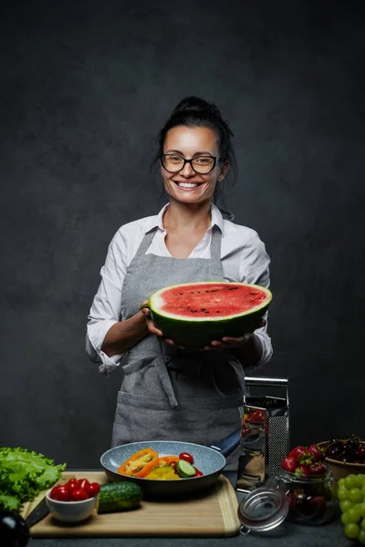 Kuchařka středního věku drží polovinu melounu v kuchyni, usmívá se a dívá se do kamery — Stock fotografie