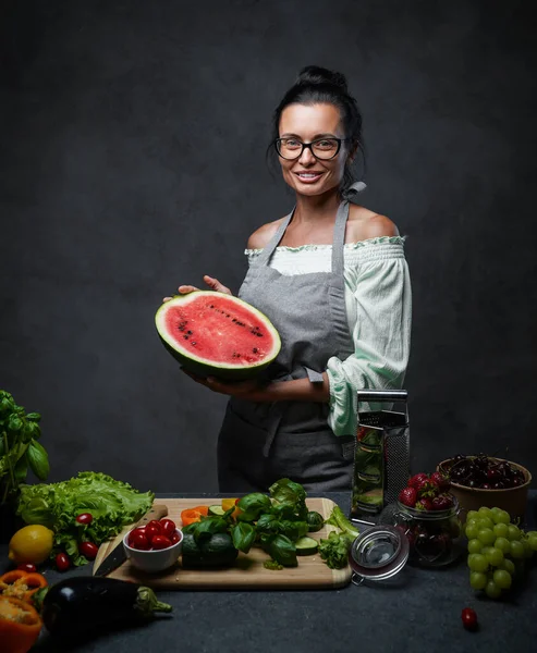 Medelålders kvinnlig kock håller hälften av vattenmelonen i köket, ler och tittar på kameran — Stockfoto