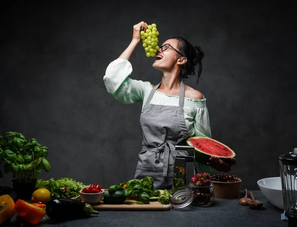 Mutfakta yemek pişiren, karpuzun yarısını tutan ve üzümleri yiyen olgun bir kadın. — Stok fotoğraf