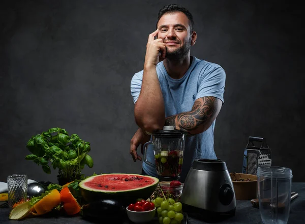 Bonito macho tatuado em camisa azul apoiando-se em um liquidificador na cozinha — Fotografia de Stock