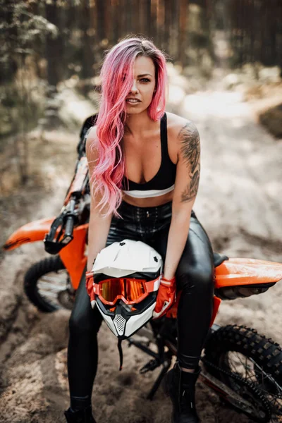 Современная татуированная девушка-хипстер с ярко-розовыми волосами, сидящая на мотокроссе в лесу — стоковое фото