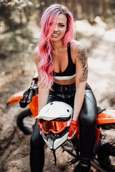 Jovem sorridente com cabelo rosa e tatuagem na mão sentada em sua moderna moto de motocross na floresta — Fotografia de Stock