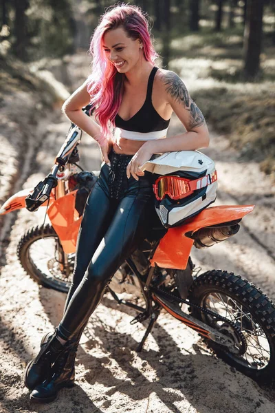 Mujer joven sonriente con cabello rosa y tatuaje en la mano sentada en su moderna moto de motocross en el bosque —  Fotos de Stock