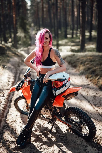 Mujer joven sonriente con cabello rosa y tatuaje en la mano sentada en su moderna moto de motocross en el bosque —  Fotos de Stock