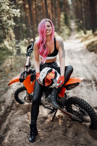Giovane donna sorridente con capelli rosa e tatuaggio a portata di mano seduta sulla sua moderna motocross bike in legno — Foto Stock