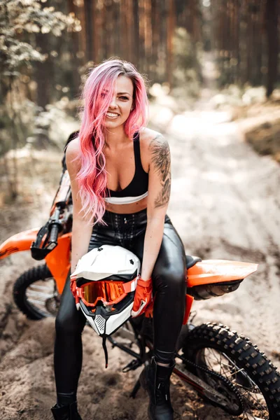 Χαμογελαστή νεαρή γυναίκα με ροζ μαλλιά και τατουάζ στο χέρι κάθεται στο σύγχρονο motocross ποδήλατό της στο δάσος — Φωτογραφία Αρχείου