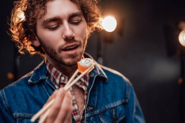 Kıvırcık saçlı, kot ceketli, sıcak suşi yiyen genç bir adamın stüdyo portresi.