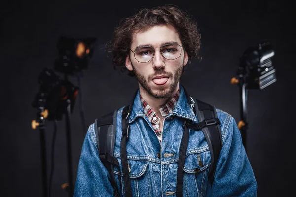 Lustiger gutaussehender Mann mit lockigem Haar trägt Jeansjacke mit Rucksack und zeigt Zunge — Stockfoto