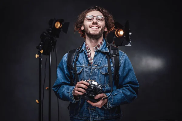 Портрет счастливого красивого кудрявого парня в очках в джинсовой куртке держит цифровую камеру в темной фотостудии — стоковое фото