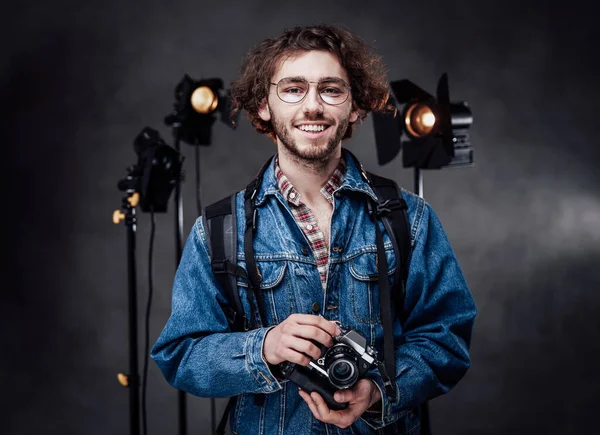 Portrait of a happy handsome curly guy in glasses wearing denim jacket holds a digital camera in dark photo studio