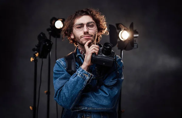 Jovem fotógrafo em óculos detém câmera digital com a mão no queixo, parece pensativo — Fotografia de Stock