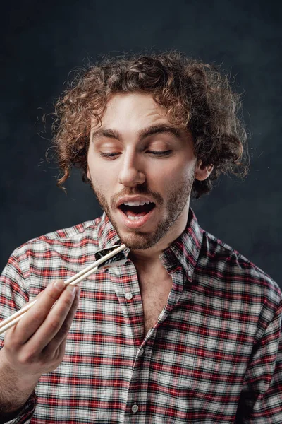 Fešák muž s kudrnatým účesem na sobě kostkované tričko jíst sushi rolky na tmavém pozadí — Stock fotografie