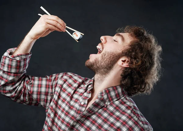 Fešák muž s kudrnatým účesem na sobě kostkované tričko jíst sushi rolky na tmavém pozadí — Stock fotografie