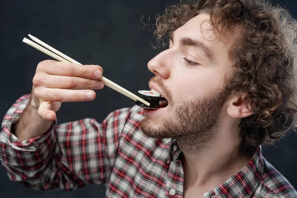 Schöner Mann mit lockiger Frisur trägt kariertes Hemd und isst Sushi-Rollen auf dunklem Hintergrund — Stockfoto