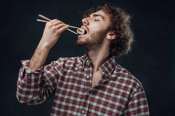 Homem bonito com penteado encaracolado vestindo camisa quadriculada comendo rolos de sushi no fundo escuro — Fotografia de Stock
