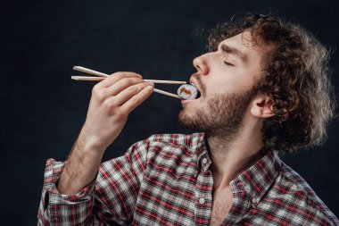 Kıvırcık saçlı, kareli gömlek giyen, koyu arkaplanda suşi yiyen yakışıklı bir adam.