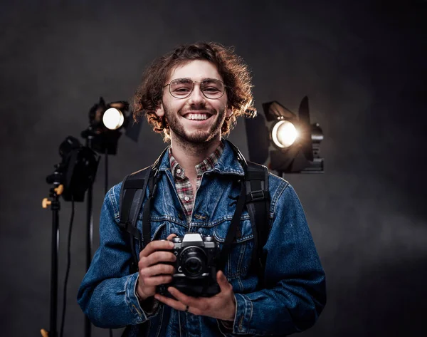 Retrato de um cara bonito feliz encaracolado em óculos usando jaqueta de ganga detém uma câmera digital no estúdio de fotos escuras — Fotografia de Stock