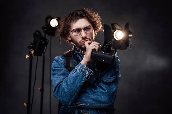 Joven fotógrafo en gafas sostiene la cámara digital con la mano en la barbilla, se ve reflexivo — Foto de Stock