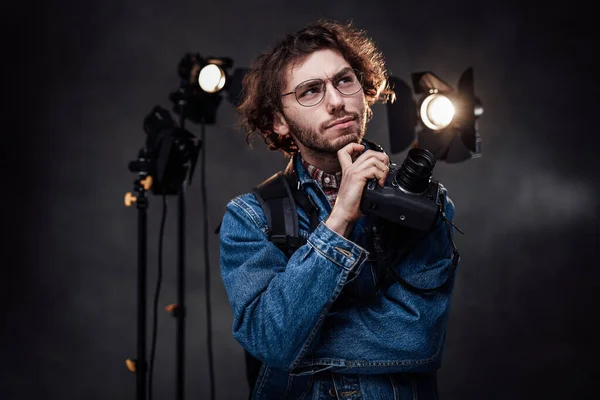 Joven fotógrafo en gafas sostiene la cámara digital con la mano en la barbilla, se ve reflexivo — Foto de Stock