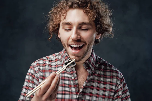 Красивый мужчина с кудрявой прической в клетчатой рубашке, поедающий суши-роллы на тёмном фоне — стоковое фото