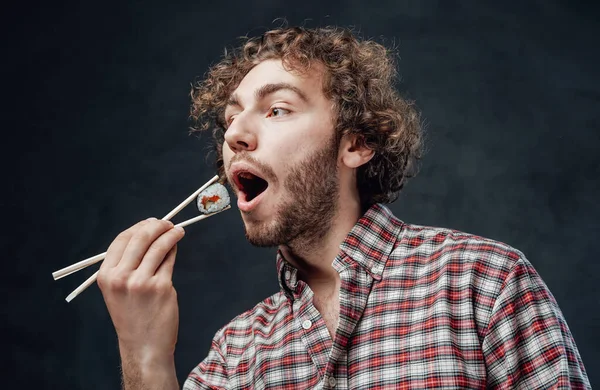 Handsome man with curly hairstyle wearing checkered shirt eating sushi rolls on dark background — Stock Photo, Image