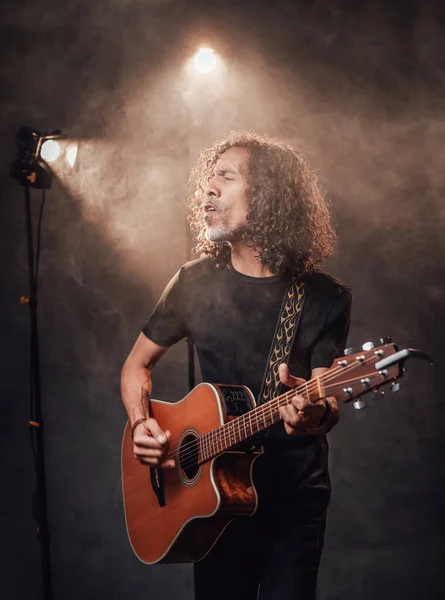 Hispanic musician emotionally singing and playing guitar in stage lights, surrounded by smoke