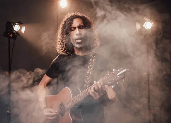 Hispanic musician emotionally singing and playing guitar in stage lights, surrounded by smoke — Stock Photo, Image