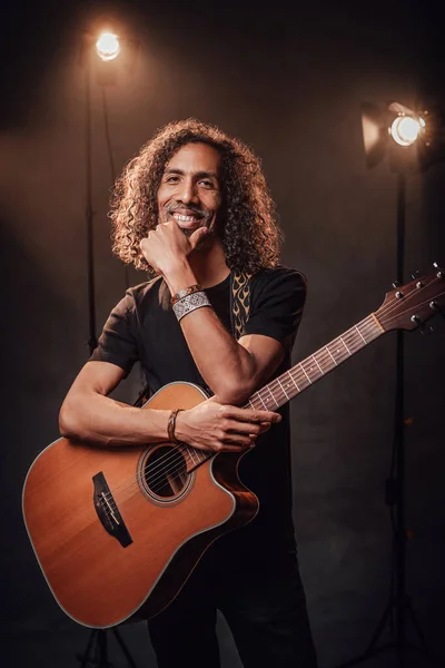 Homem hispânico de meia-idade músico de t-shirt preta segura guitarra em uma cena, sorrindo e olhando para a câmera — Fotografia de Stock