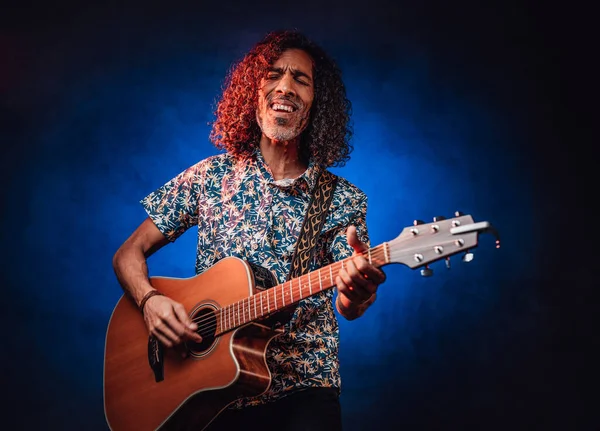 Músico hispânico emocionalmente cantando e tocando guitarra em um escuro iluminado por luz azul e vermelha — Fotografia de Stock