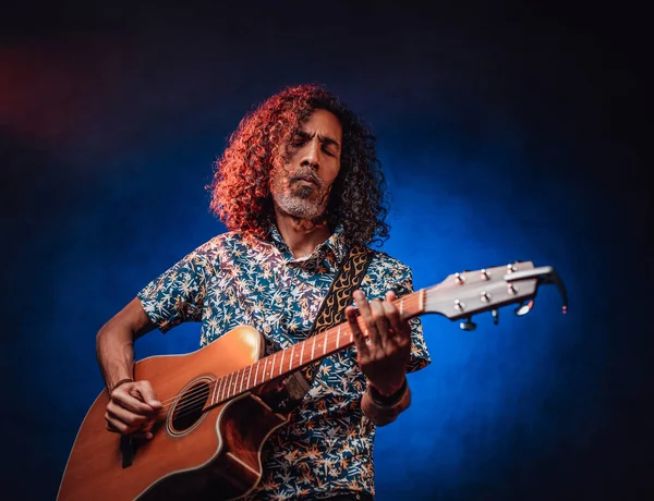 Middle aged hispanic man musician playing guitar on a dark illuminated by blue and red light — Stock Photo, Image