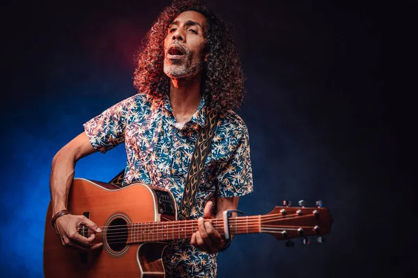 Músico hispânico emocionalmente cantando e tocando guitarra em um escuro iluminado por luz azul e vermelha — Fotografia de Stock