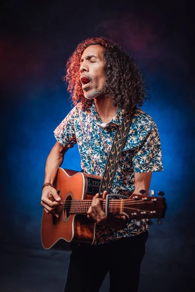 Hispanic musician emotionally singing and playing guitar on a dark illuminated by blue and red light — Stock Photo, Image
