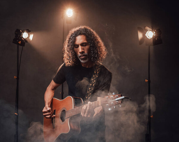 Middle aged hispanic musician emotionally playing guitar in stage lights, surrounded by smoke