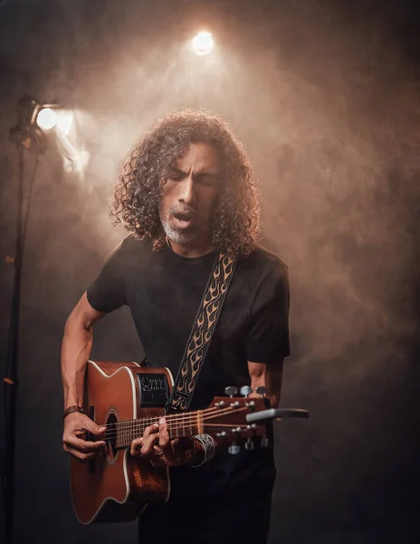 Hispanic musician emotionally singing and playing guitar in stage lights, surrounded by smoke — Stock Photo, Image