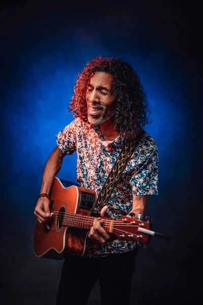 Hispanic musician emotionally singing and playing guitar on a dark illuminated by blue and red light — Stock Photo, Image