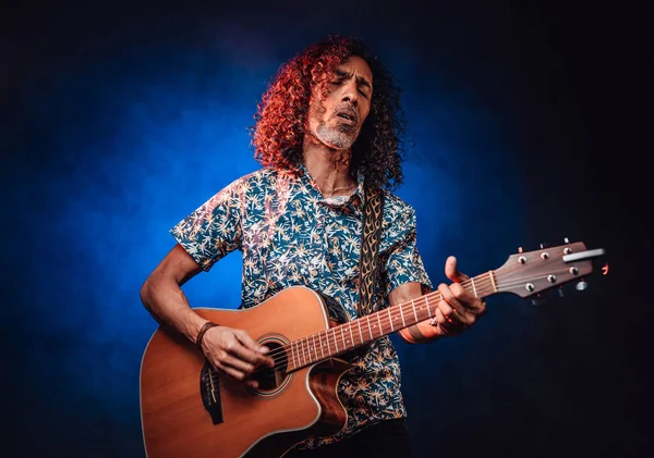 Medelålders latinamerikan musiker spelar gitarr på en mörk upplyst av blått och rött ljus — Stockfoto