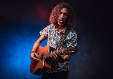 Orta yaşlı İspanyol müzisyen. Mavi ve kırmızı ışıkla aydınlatılmış koyu bir ışıkta gitar çalıyor.