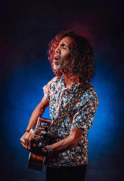 Hispanic musician emotionally singing and playing guitar on a dark illuminated by blue and red light — Stock Photo, Image