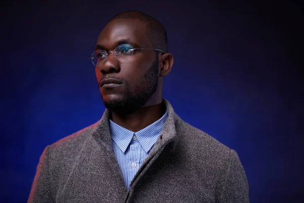 Stylish African American man in gray jacket and glasses on dark background with blue light — Stock Photo, Image