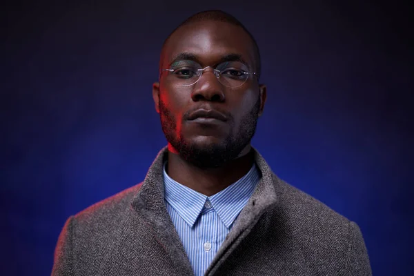 Stylish African American man in gray jacket and glasses on dark background with blue light — Stock Photo, Image
