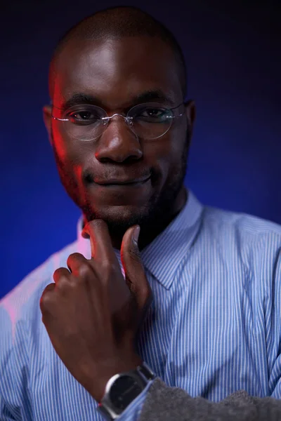 Africký americký student v modré košili a brýlích, drží ruku na bradě a usmívá se — Stock fotografie
