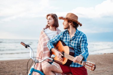 Bisikletli bir çift. Oğlan gitarda çalıyor. Kız pembe battaniyeye sarılmış, gün batımını seyrediyor.