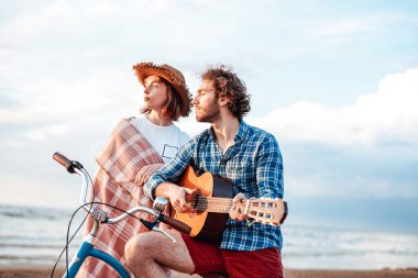 Oğlan gitarda oynuyor, mavi bisikletin üzerinde oturuyor. Kız arkasında duruyor.