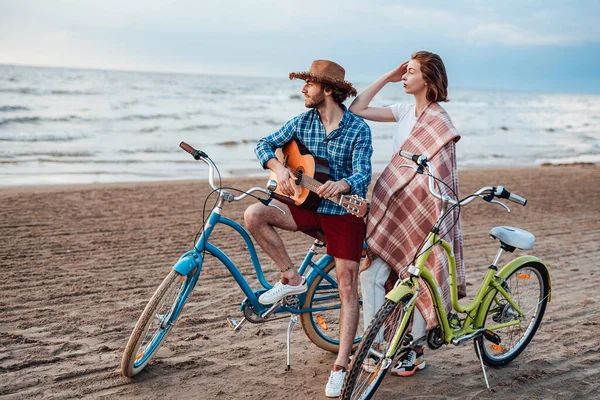 Två personer står med sina cyklar. Pojken spelar på gitarr, i fin hatt. Flickan är insvept i rosa filt — Stockfoto