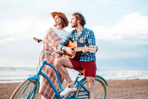 Unga par med sin cykel. Pojken spelar på gitarr. Flickan är insvept i en rosa filt, tittar på solnedgången — Stockfoto