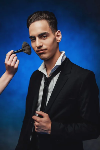 Mano del maquillador sostiene cepillo y prepara guapo elegante vestido chico para una sesión de fotos —  Fotos de Stock